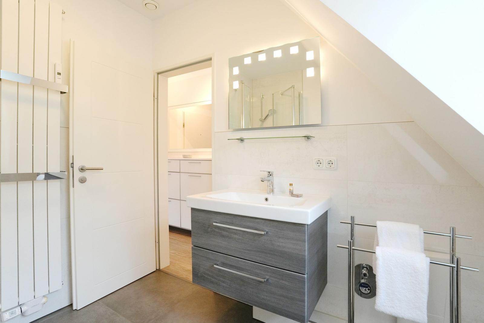 Badezimmer mit Waschbecken, Spiegel und Handtuchhalter. Tür führt in einen weiteren Raum.