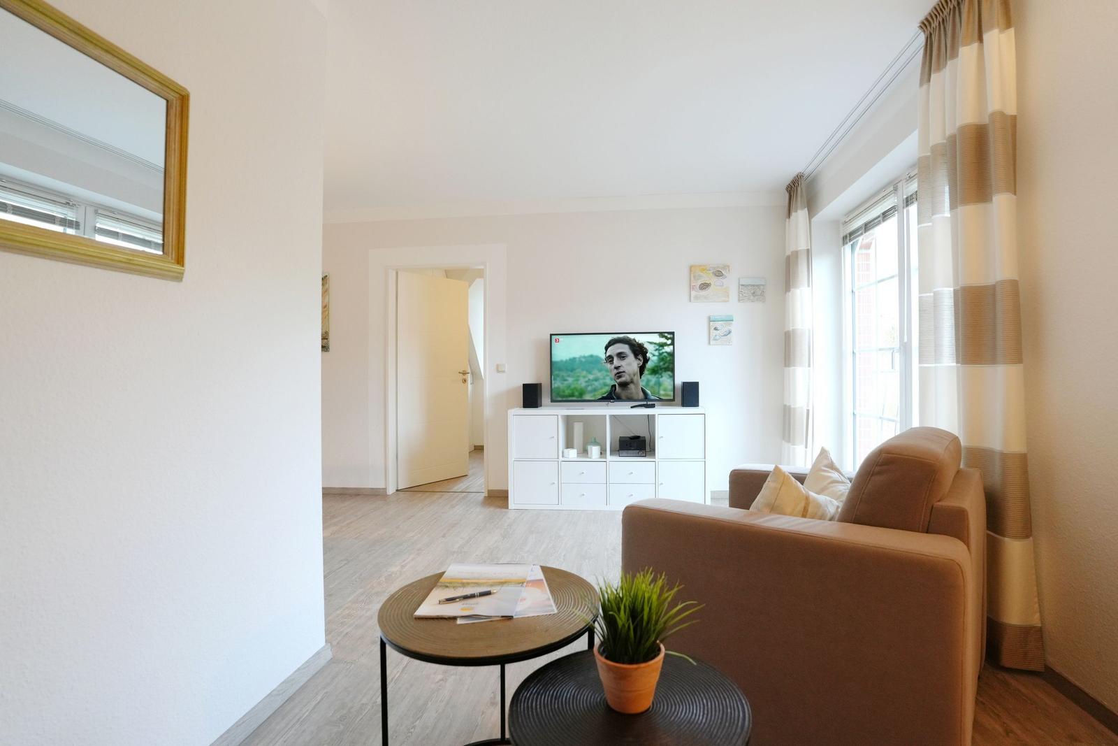 Wohnzimmer mit Sofa, Fernseher und Fenster. Holzboden und weiße Wände.