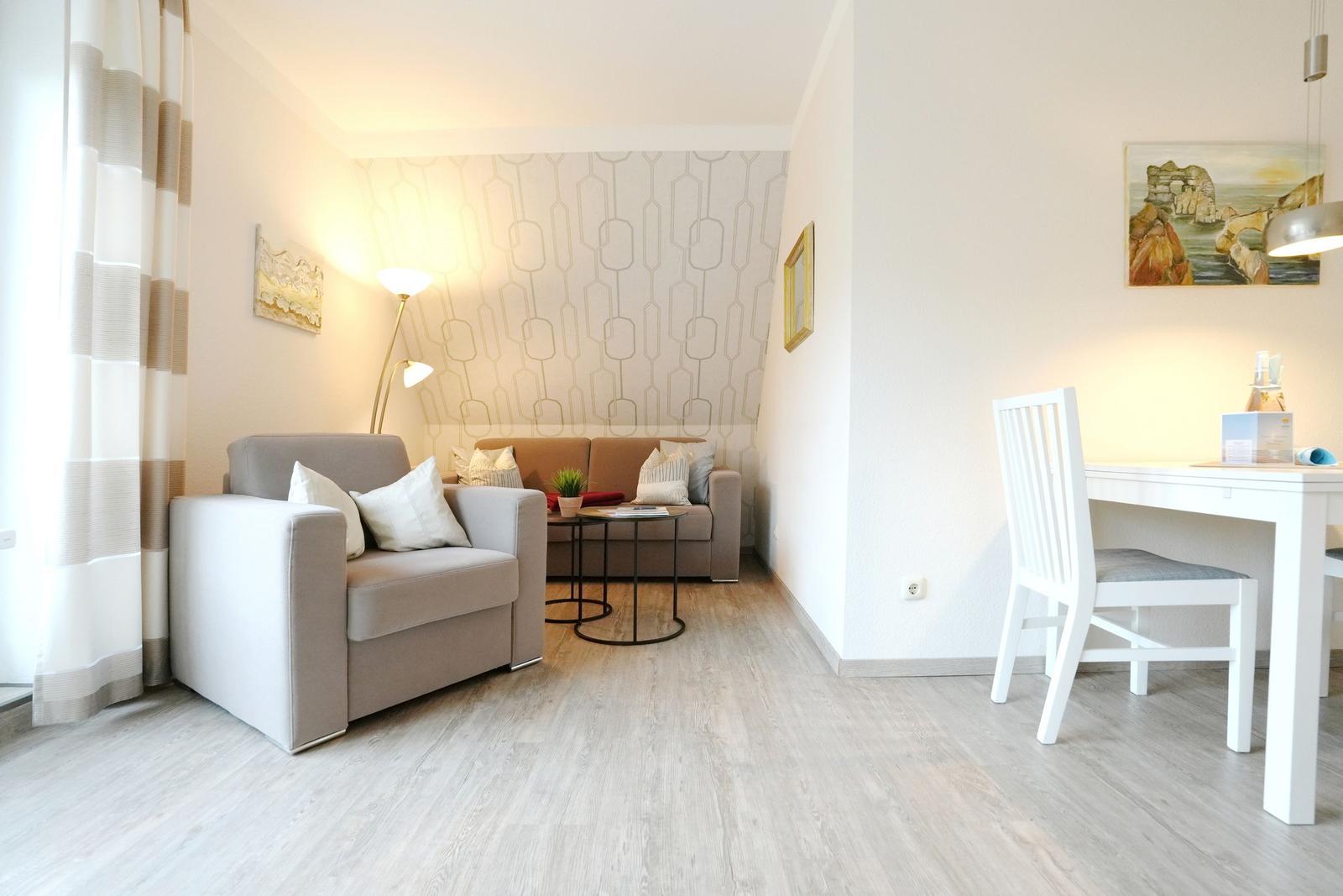 Wohn- und Essbereich mit Sofa, Sessel und Tisch. Wand mit Muster. Fenster mit Vorhängen.