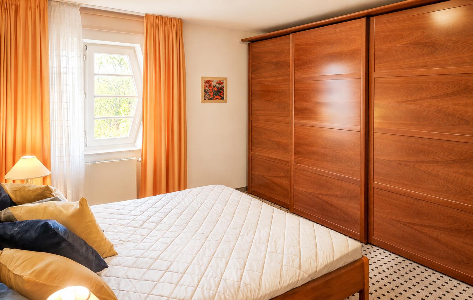 Schlafzimmer mit Bett, Holzschrank und Fenster mit orangefarbenen Vorhängen.