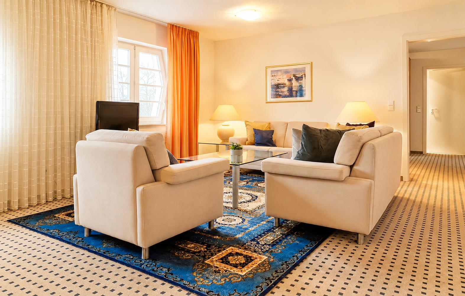 Wohnzimmer mit zwei Sesseln, Sofa, Glas-Tisch und blauem Teppich.