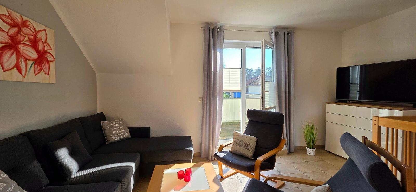 Wohnzimmer mit Sofa, Sessel und TV. Fenster mit Blick auf Balkon.