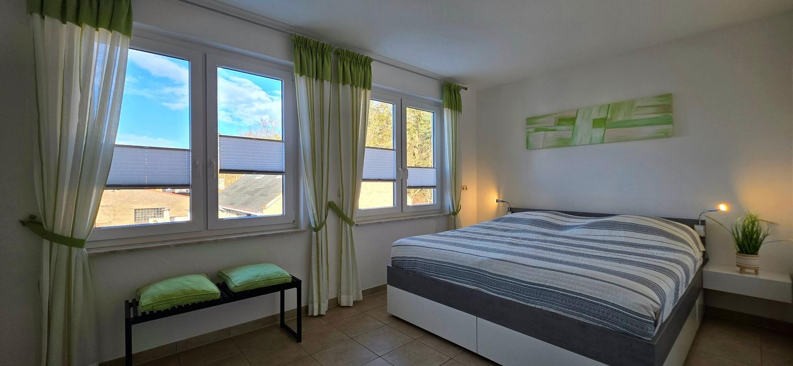 Schlafzimmer mit Bett, Fenstern und Wandbild.