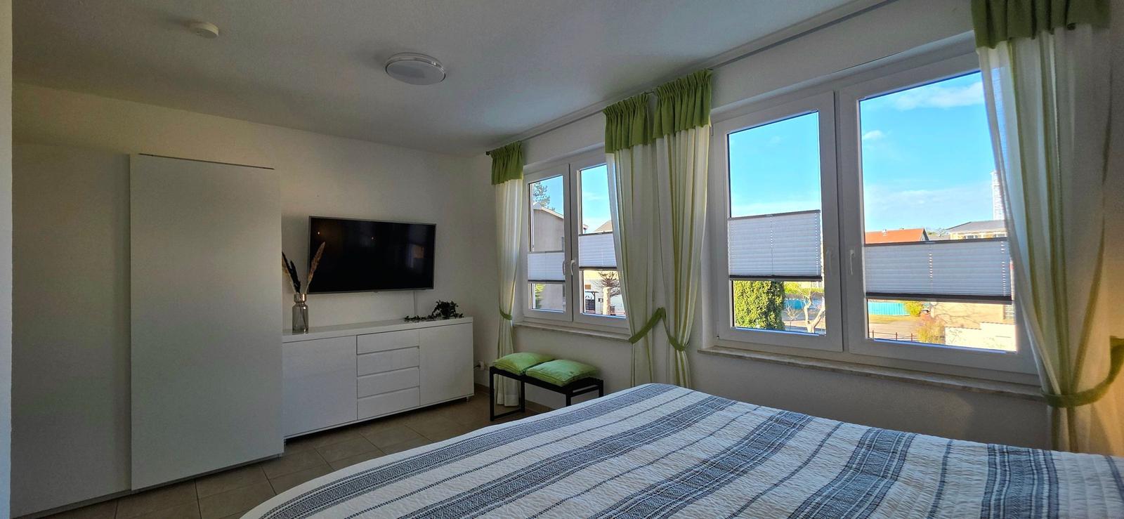 Schlafzimmer mit Bett, TV und Fenstern mit Blick auf Außenbereich.