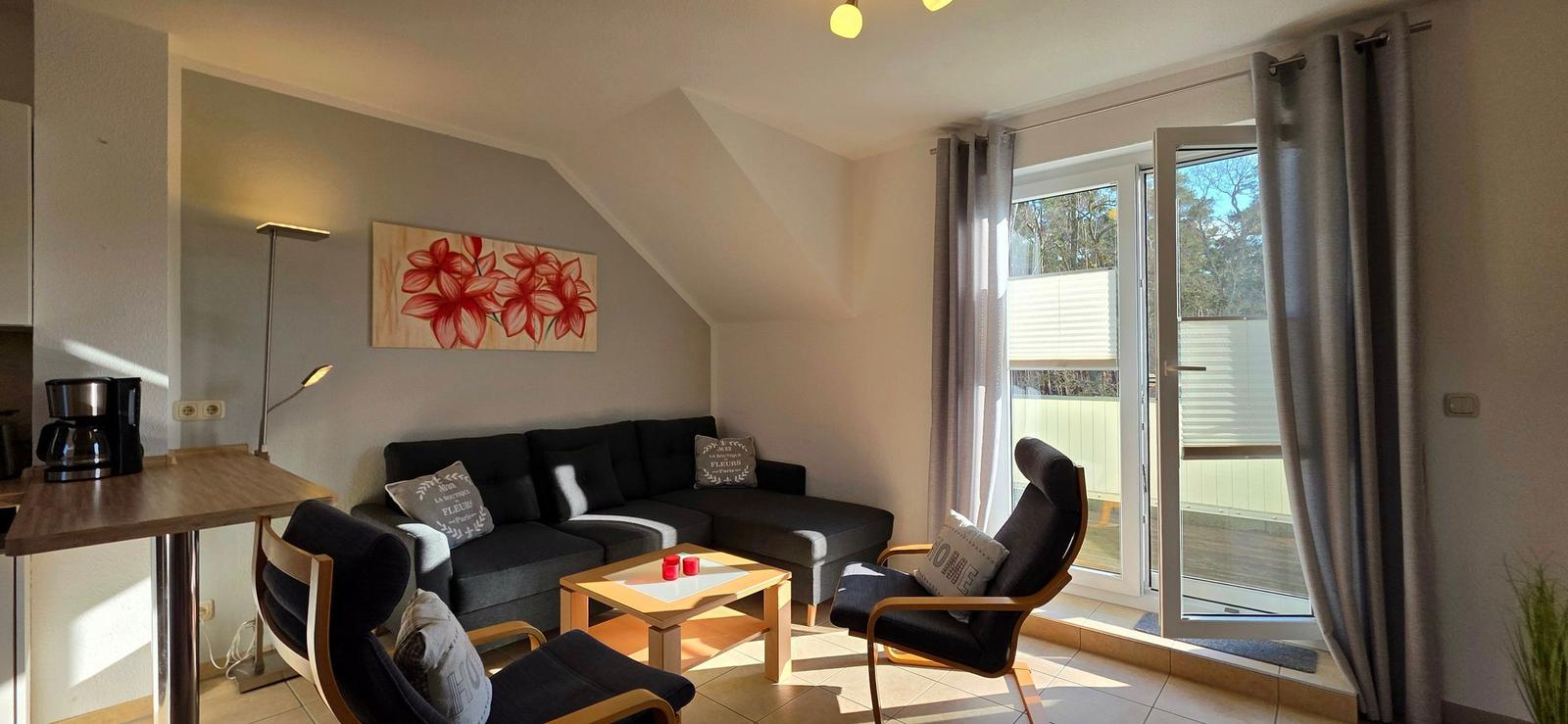 Wohnbereich mit Sofa, zwei Sesseln und Tisch. Große Glastür führt auf Balkon. Wandbild mit roten Blumen.