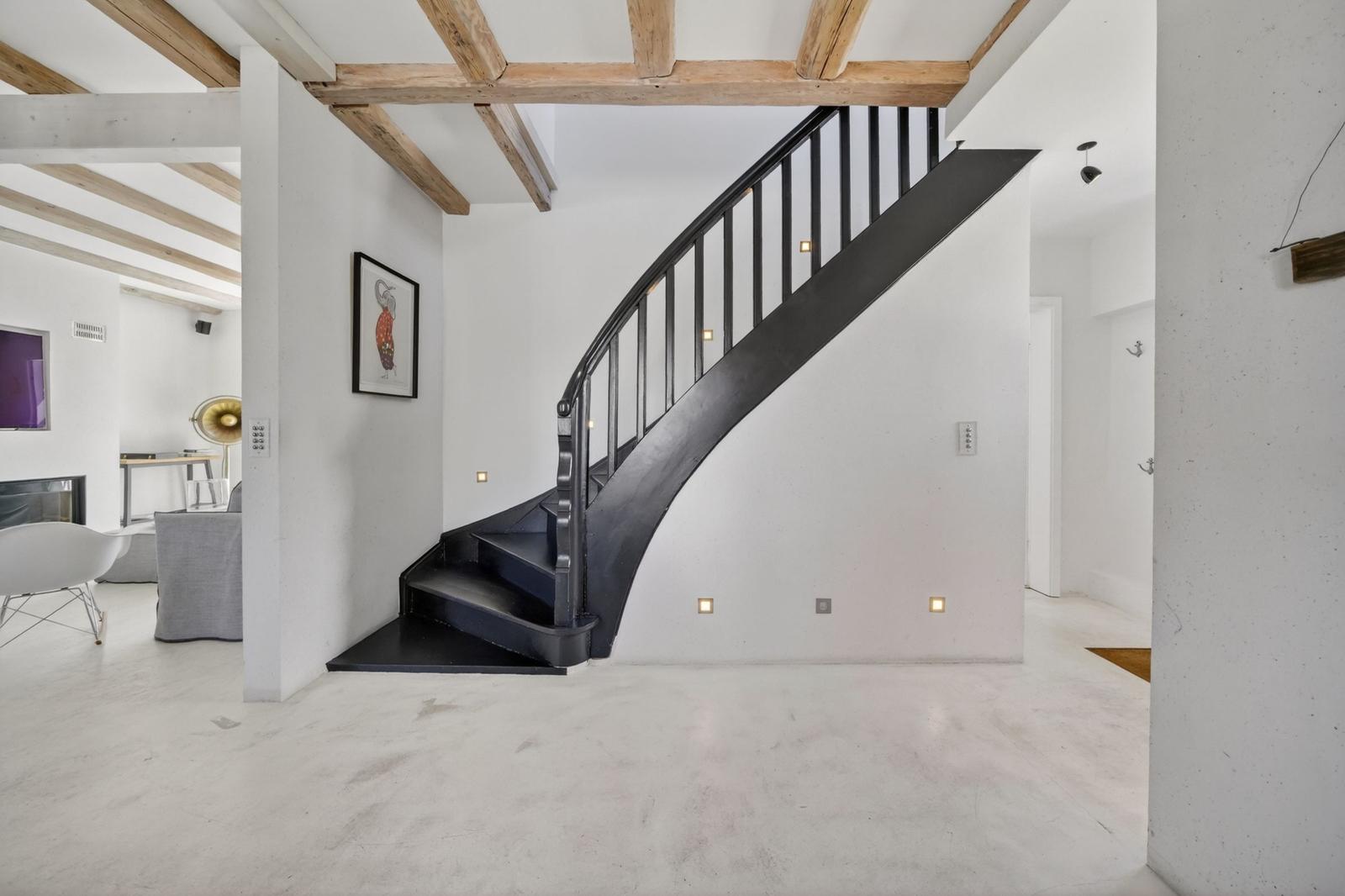 Eine Treppe mit schwarzem Geländer führt nach oben. Holzbalken und weiße Wände sind sichtbar.