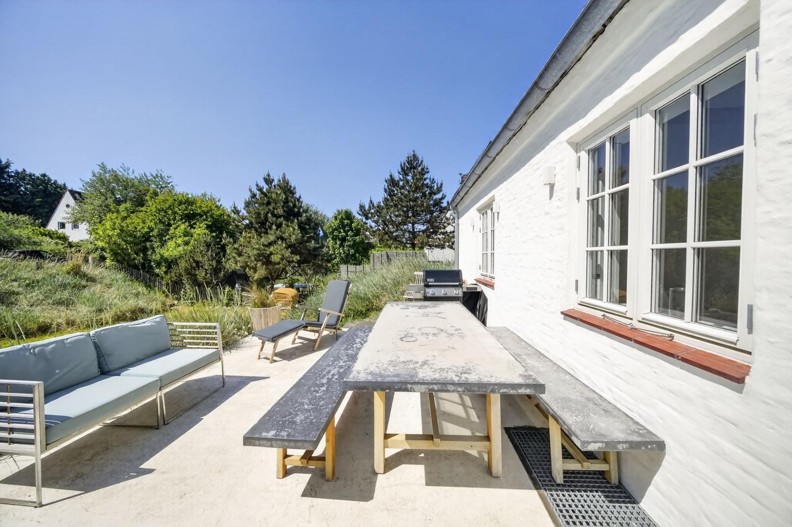 Außenbereich mit Tisch, Bänken, Sofa und Grill neben weißem Haus.