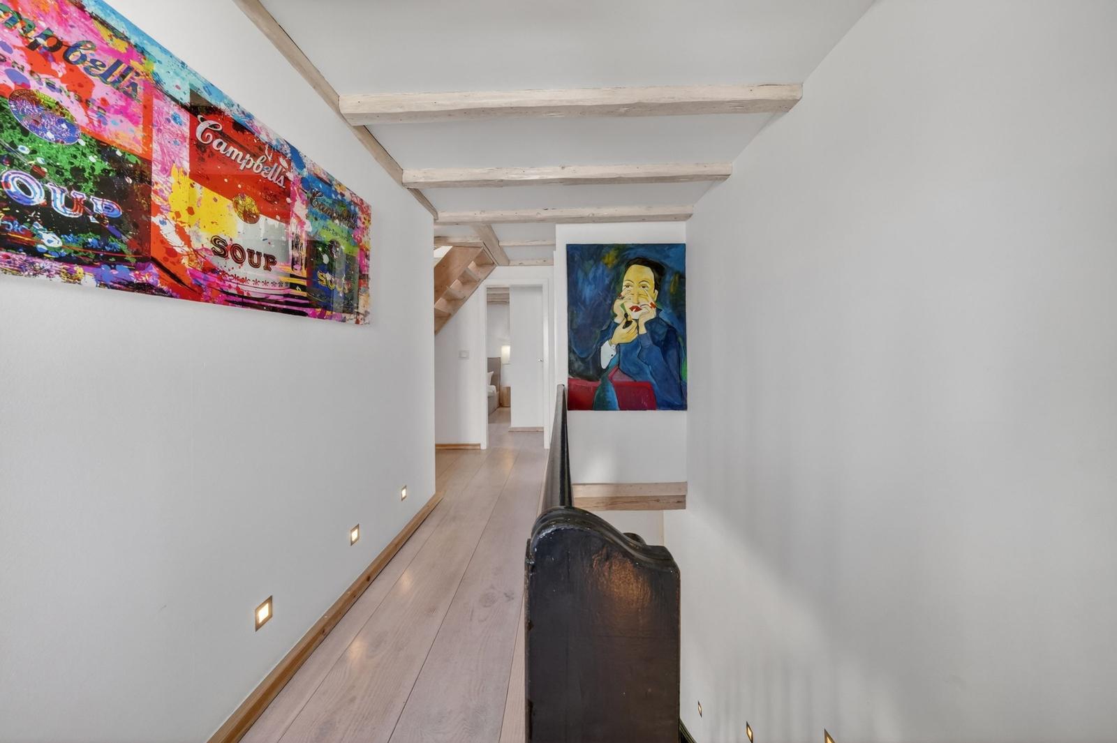 Flur mit Holzboden, Kunstwerken an den Wänden und Sicht auf Treppenbereich.