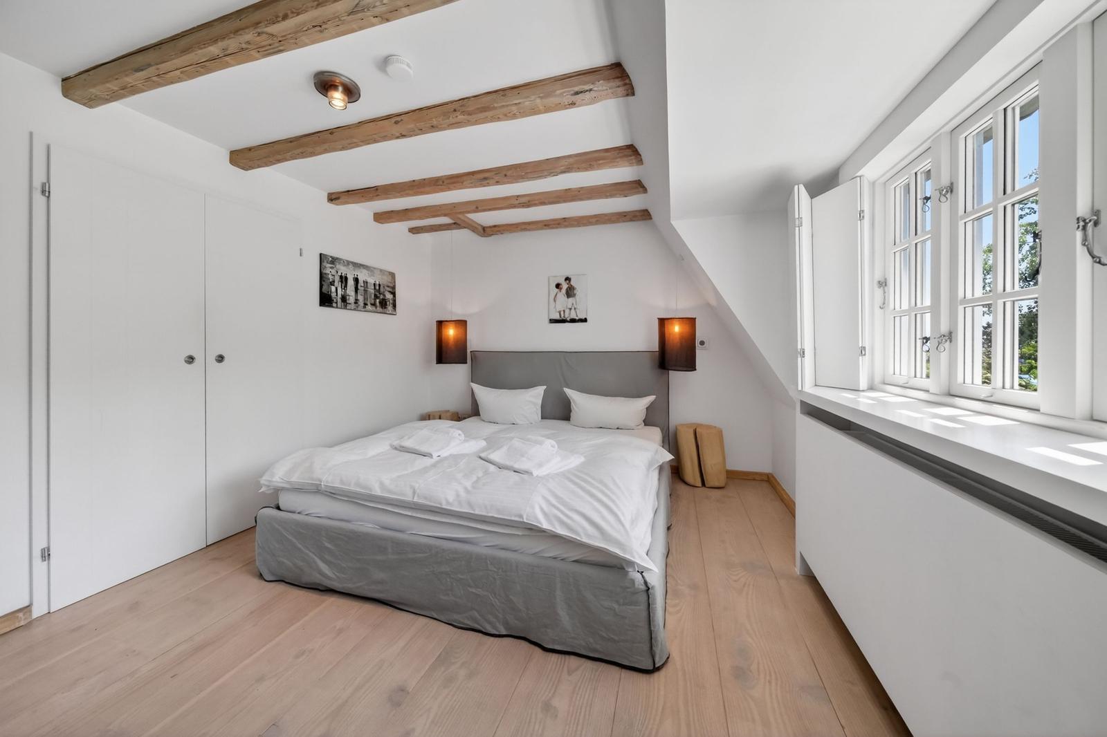 Schlafzimmer mit Dachschrägen, Holzbalken, großem Fenster und Bett mit weißer Bettwäsche.