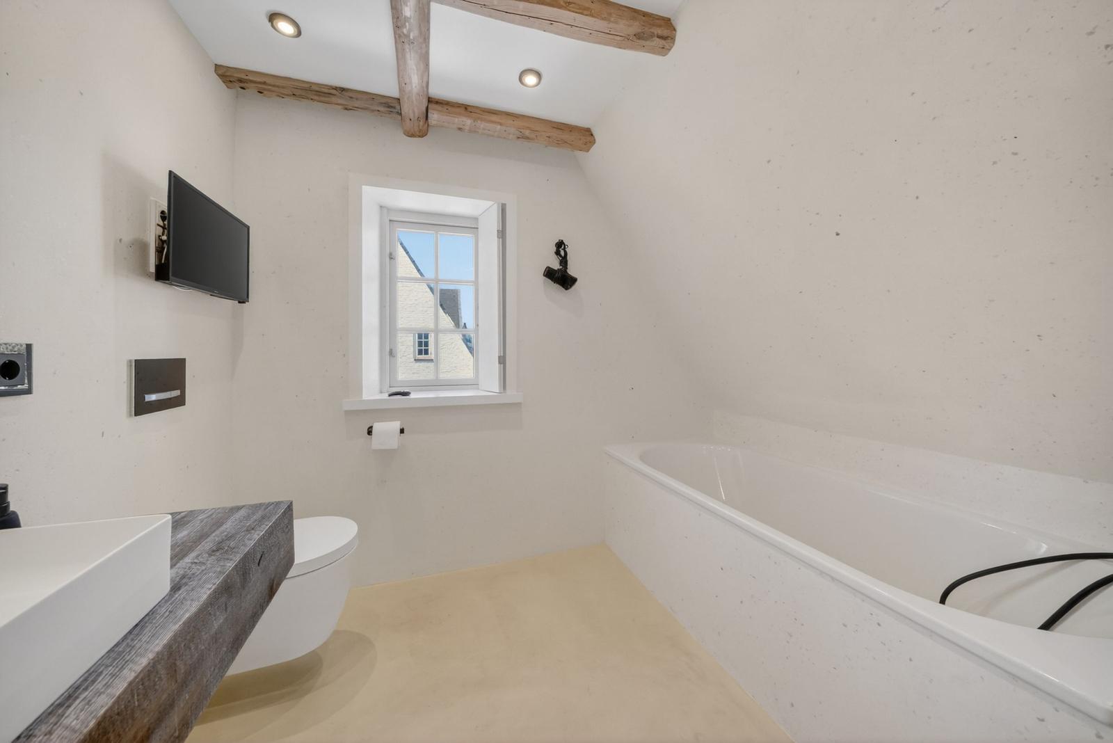 Badezimmer mit Badewanne, Toilette und Fenster. Holzbalken an der Decke.