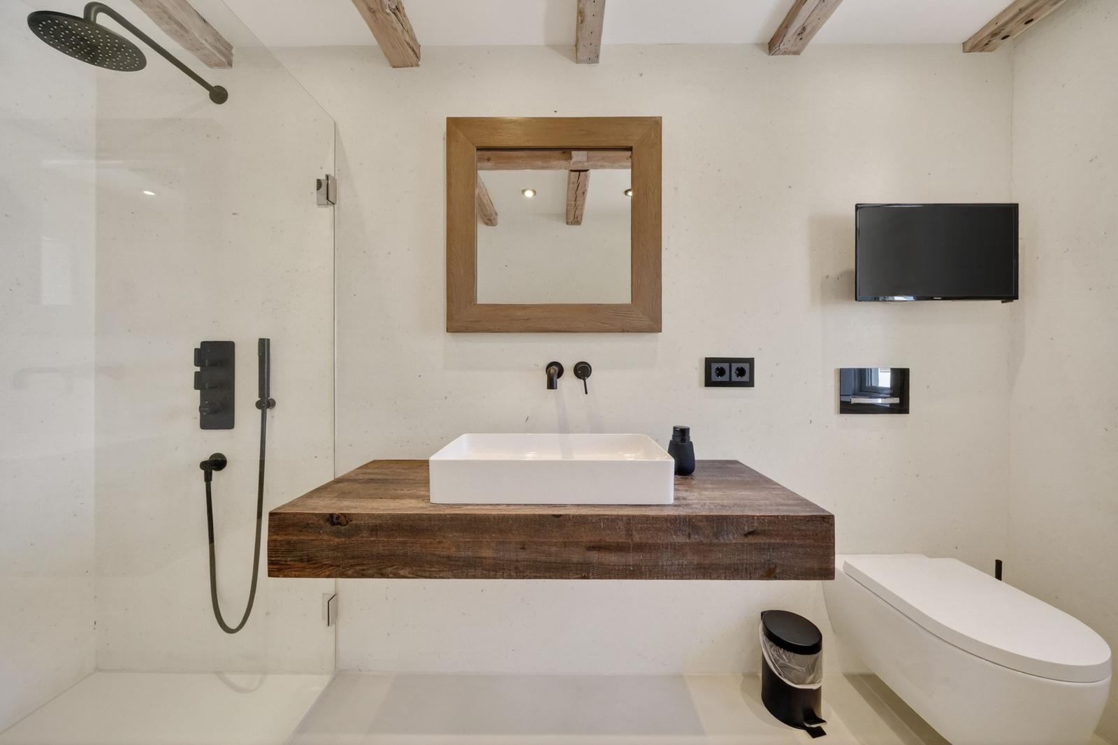 Badezimmer mit Dusche, Waschtisch auf Holzplatte und Toilette. Spiegel und TV an der Wand.