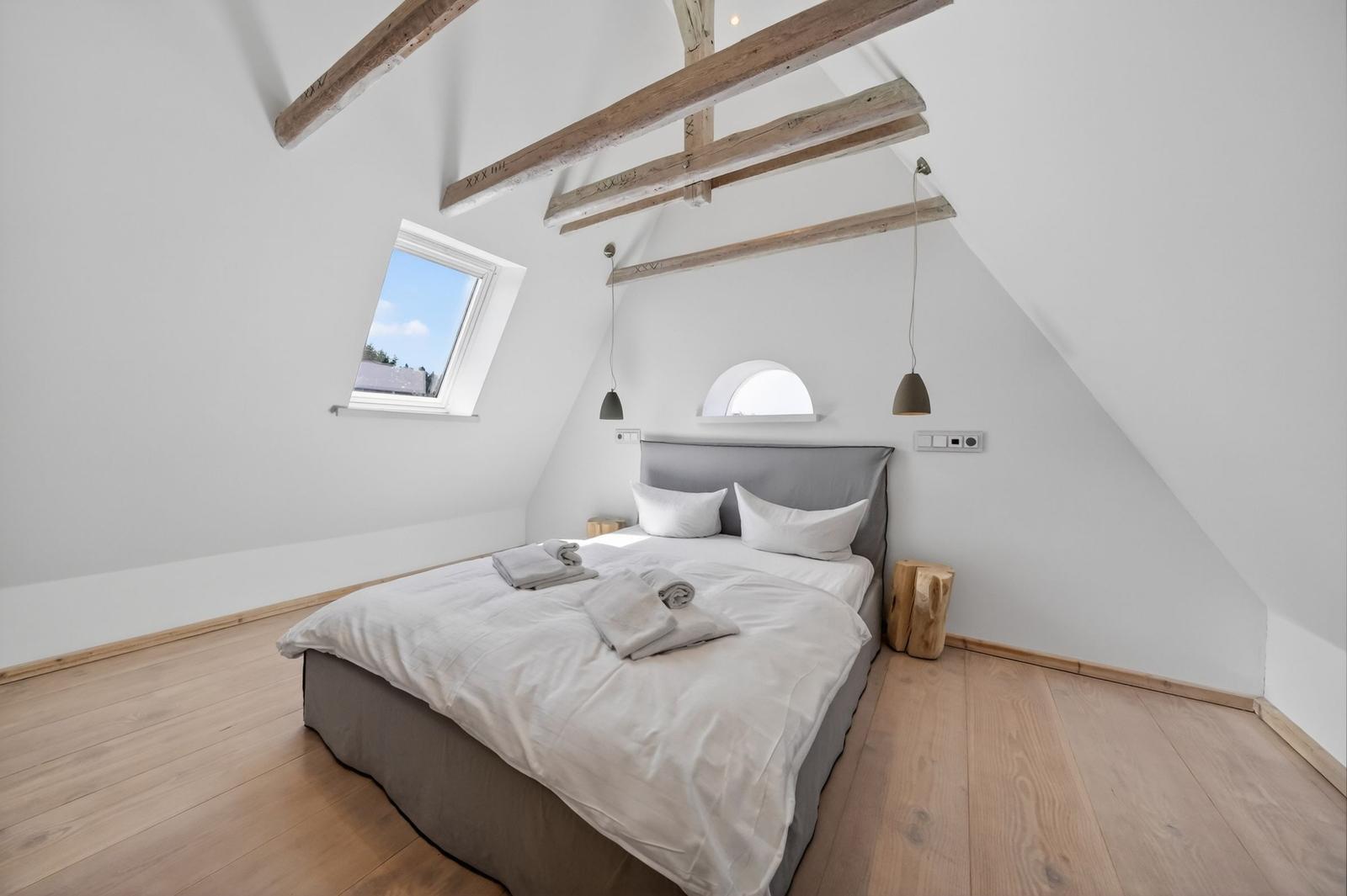 Schlafzimmer mit Dachschrägen, Holzboden und Dachfenster. Bett mit weißen Kissen und Bettwäsche.