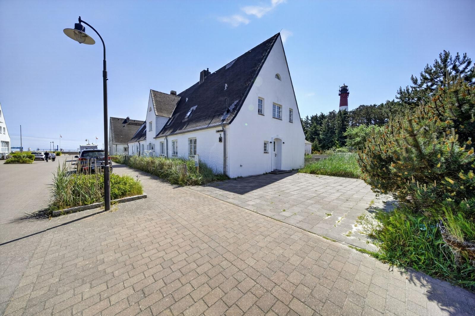 Weißes Haus mit dunklem Dach, gepflastertes Vorfeld und Leuchtturm im Hintergrund.