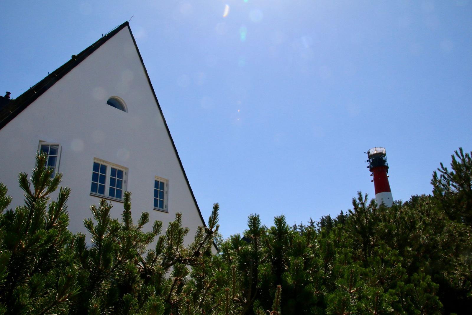 Weißes Haus mit Dach und Fenstern, umgeben von Nadelbäumen. Im Hintergrund steht ein roter Leuchtturm.