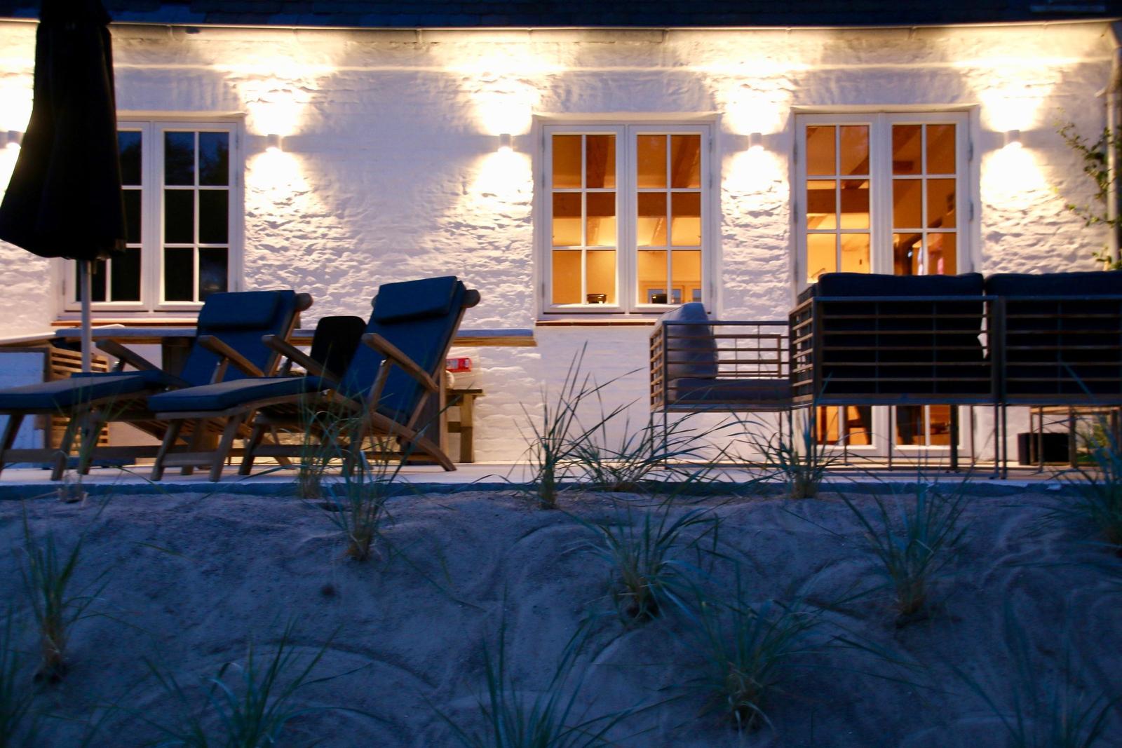 Eine Terrasse mit Liegestühlen und einem Sandbeet vor einem hellen Haus mit beleuchteten Fenstern.