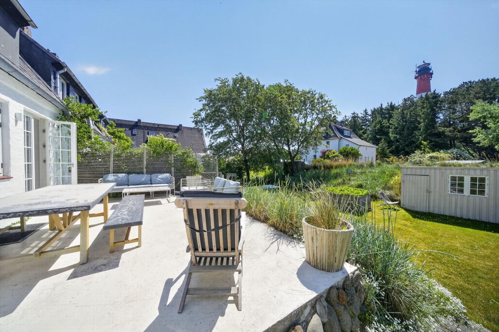 Terrasse mit Tisch, Bank und Stuhl. Grünfläche und Leuchtturm im Hintergrund.