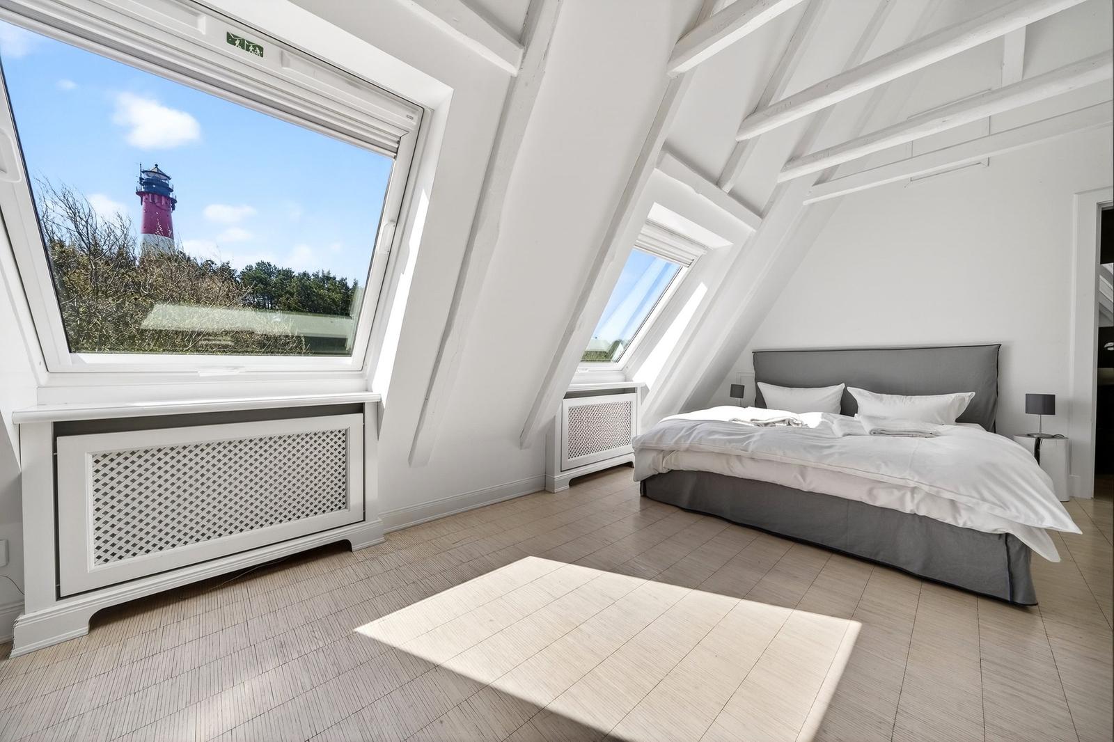 Schlafzimmer mit Blick auf Leuchtturm, weißer Balkenrahmen, graues Bett und Holzboden.