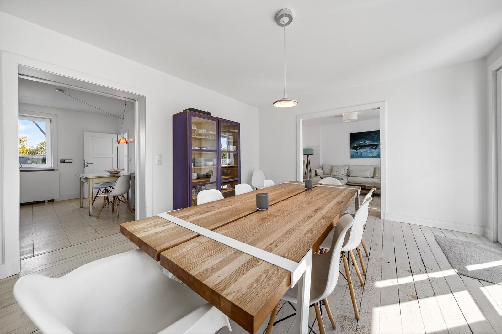 Essbereich mit langer Holztafel und weißen Stühlen. Durchgänge zu Wohn- und Küchenbereich.