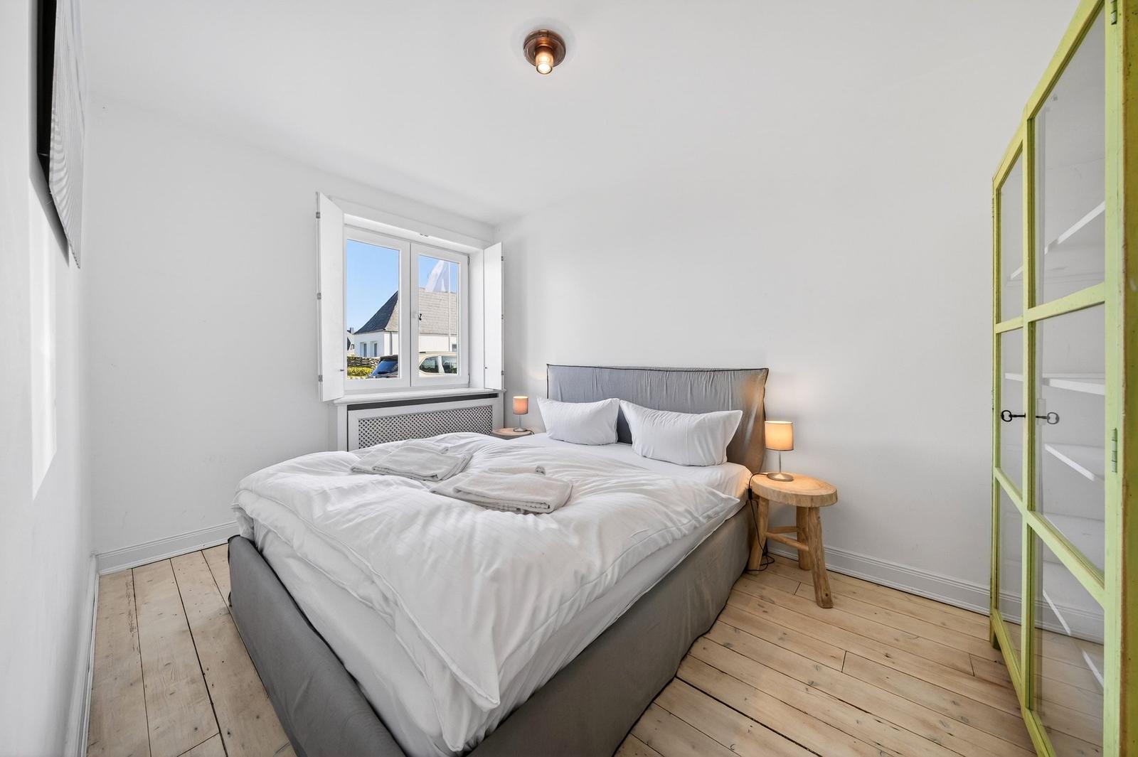 Schlafzimmer mit Bett, Nachttisch und Fenster. Holzboden und Kleiderschrank.