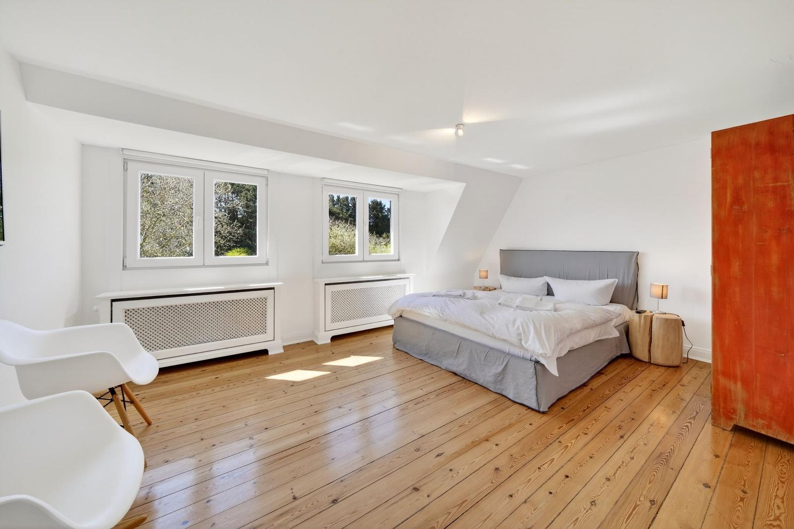Schlafzimmer mit Holzboden, Bett, Fenstern und weißen Wänden.