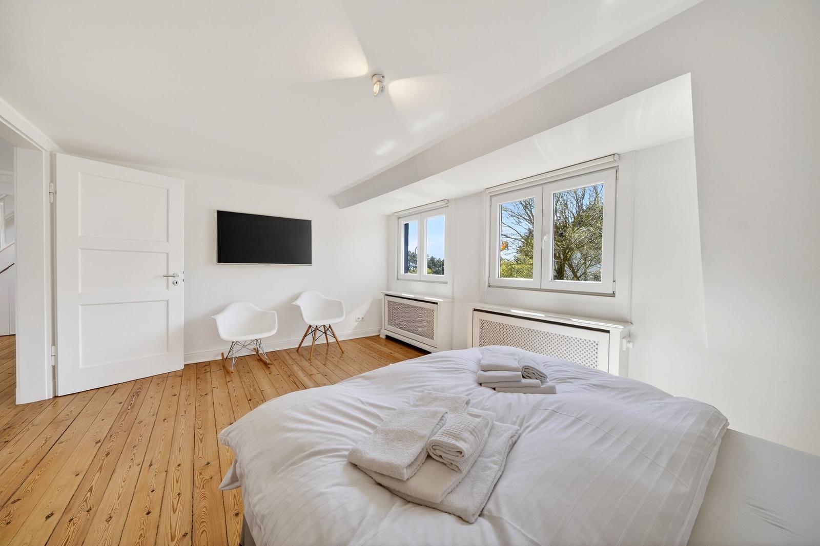 Schlafzimmer mit Bett, zwei Fenstern und Holzboden. Fernseher an der Wand.