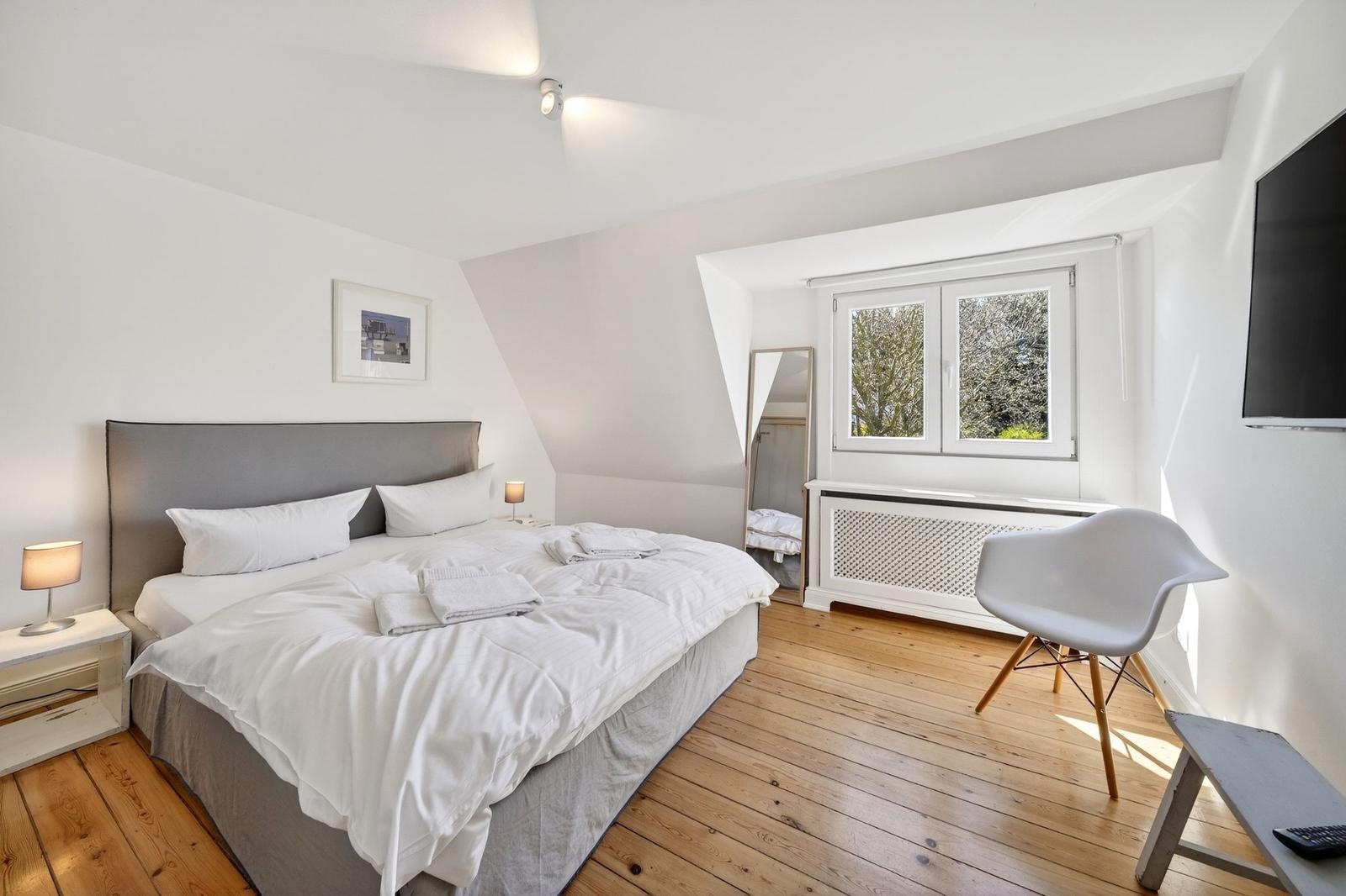 Schlafzimmer mit Doppelbett, Fenster und Stuhl auf Holzboden.