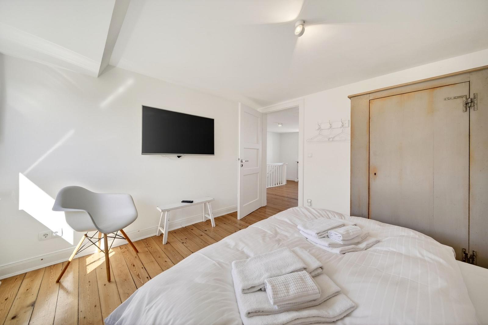 Schlafzimmer mit Bett, Fernseher und Stuhl auf Holzboden.