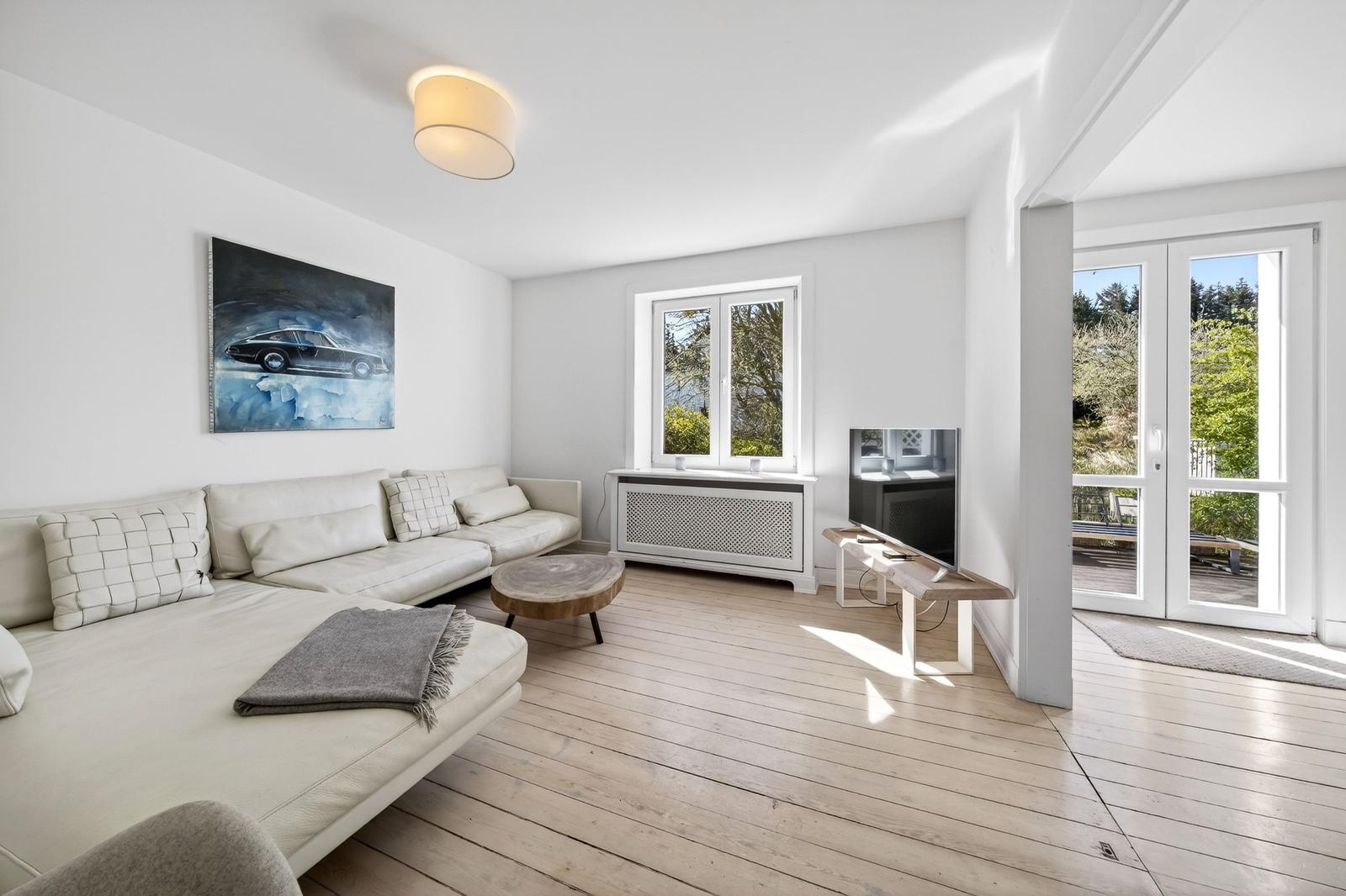 Wohnzimmer mit Sofa, Couchtisch und Fernseher. Durchgang zur Terrasse mit Blick auf Grünflächen.