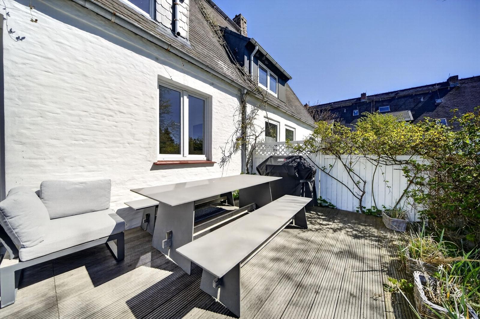 Terrasse mit Sitzbank, Tisch und Grill. Weiße Wand und Holzdecke. Grünzeug im Hintergrund.