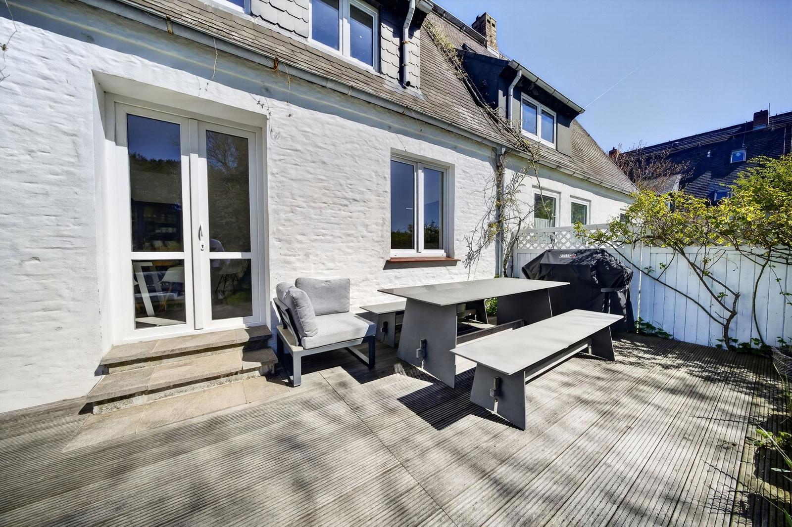 Terrasse mit Sitzgruppe, Tisch und Grill. Weiße Hauswand und Holzdecke.