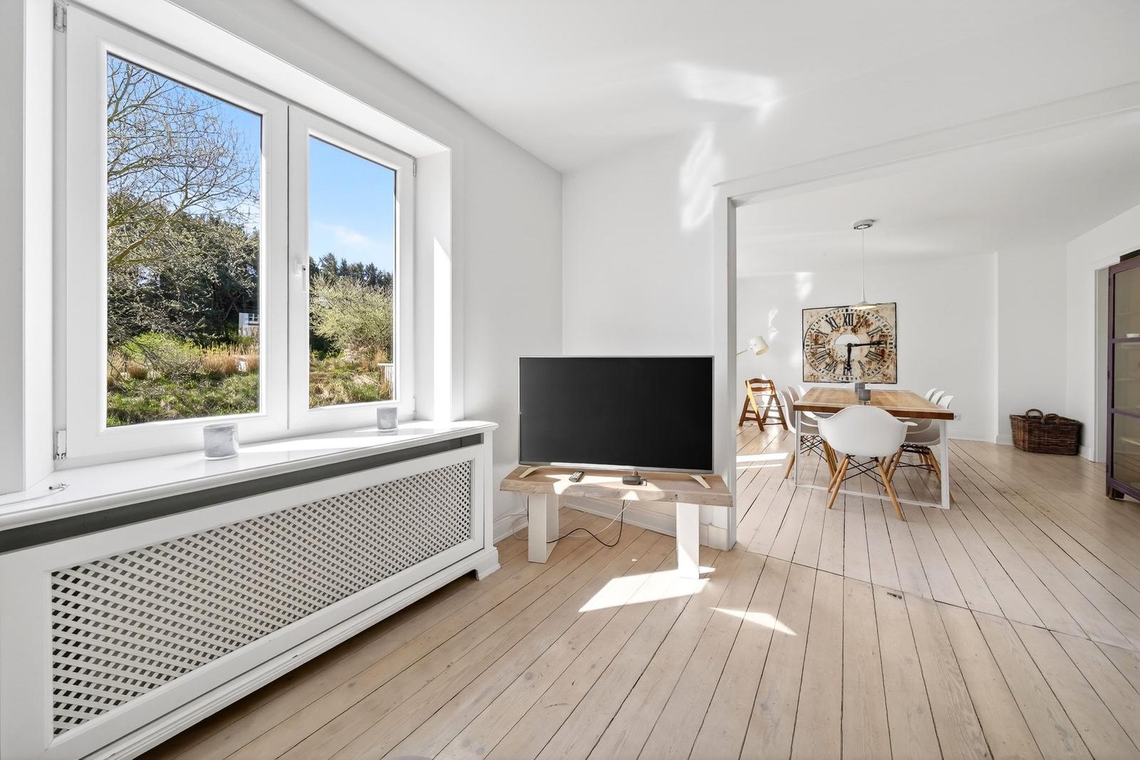 Wohnzimmer mit Fernseher und Blick auf Garten. Essbereich mit Tisch und Stühlen sichtbar.
