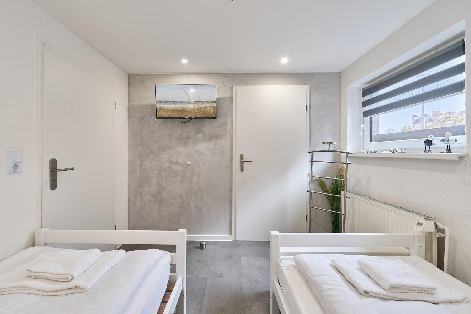 Zwei Betten mit weißen Laken, Fernseher an der Wand, Fenster mit Rollos.