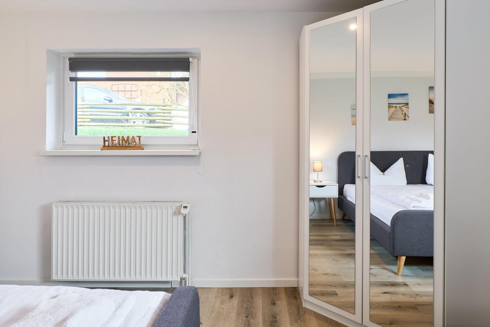 Schlafzimmer mit Spiegelschrank, Fenster und Heizung. Bett im Spiegel sichtbar.