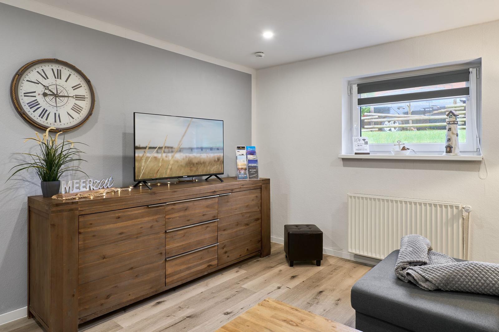 Wohnzimmer mit Fernseher, Holzschrank und Fensterblick auf Grünfläche.