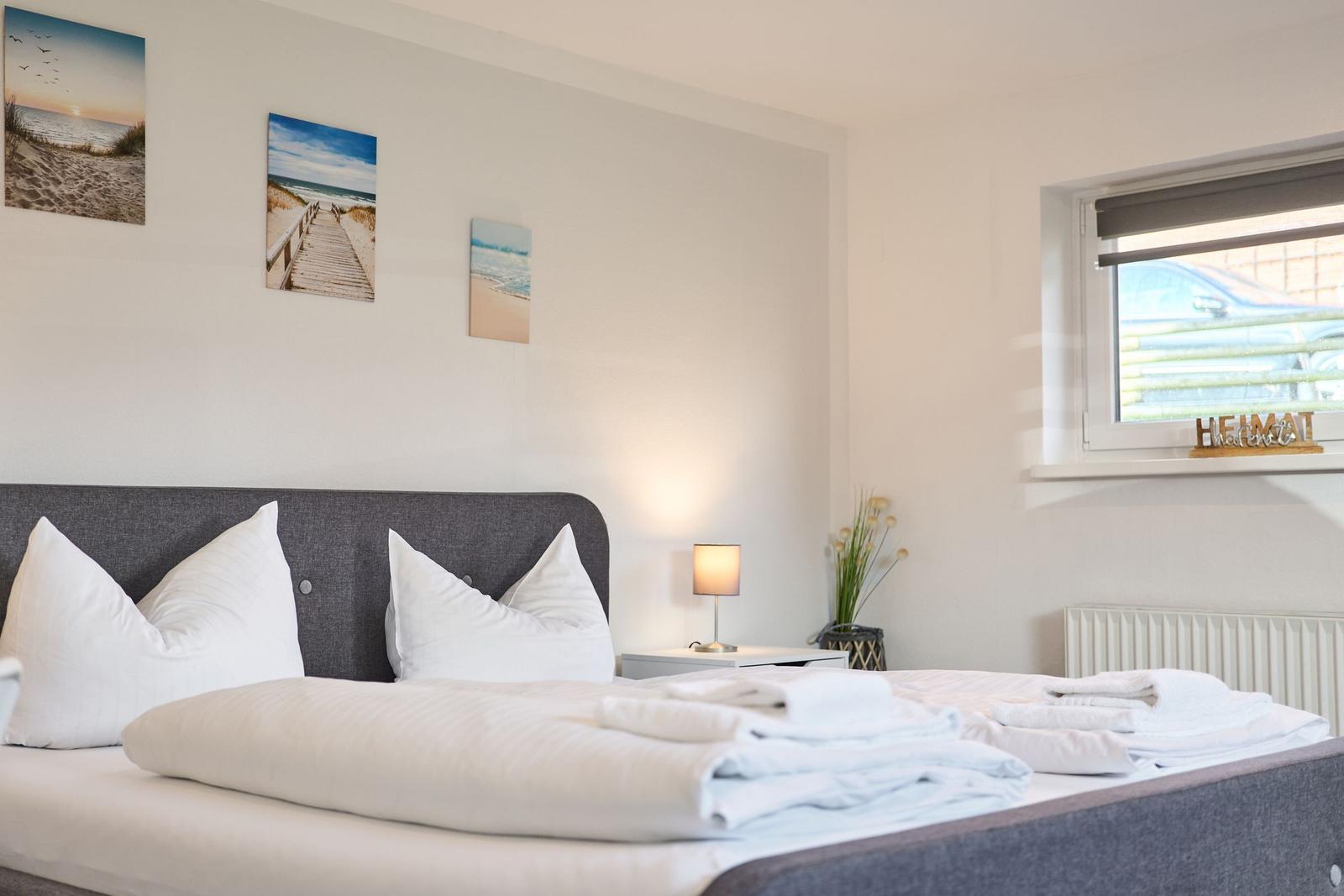 Schlafzimmer mit Bett, Nachttisch und Fenster. Wandbilder zeigen Strand.