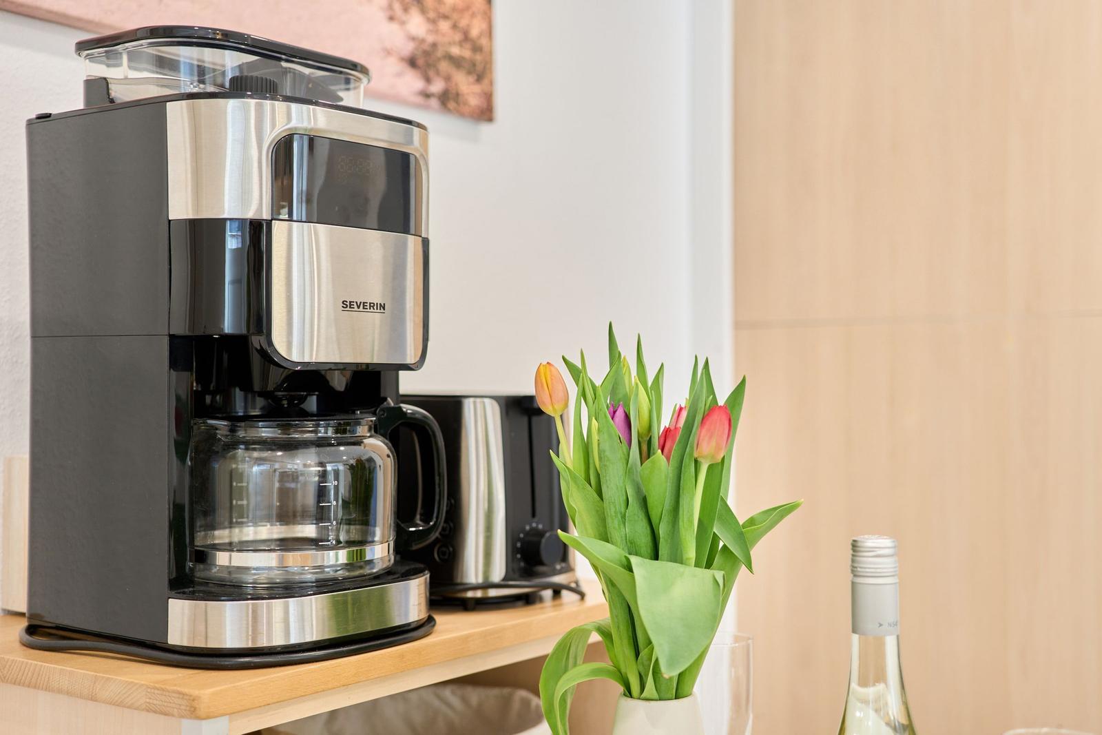 Eine Kaffeemaschine steht auf einer Arbeitsplatte neben einer Tulpenblume und einer Weinflasche.