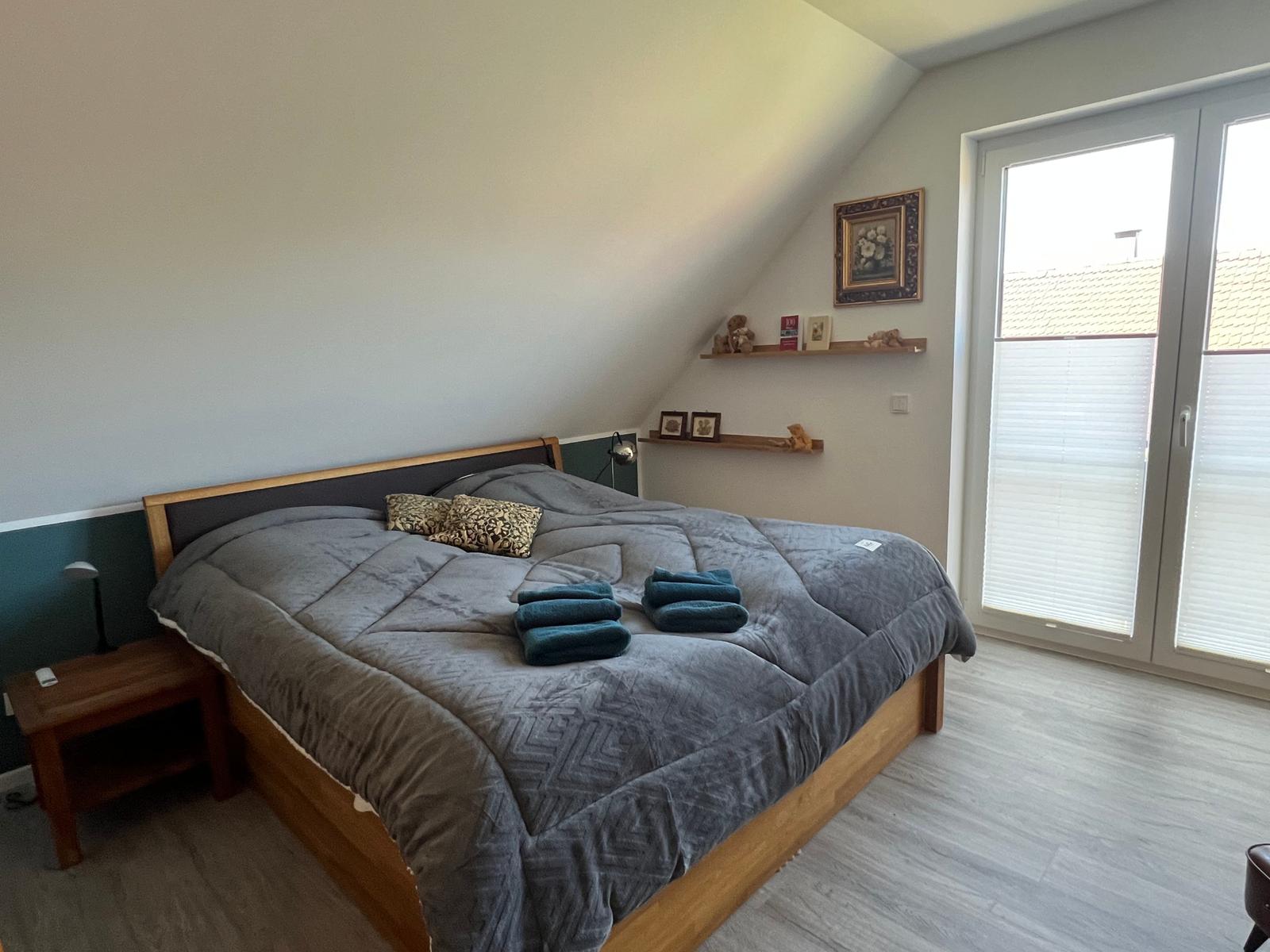 Schlafzimmer mit Dachschräge, Bett, Nachttisch und Fenster mit Jalousien.