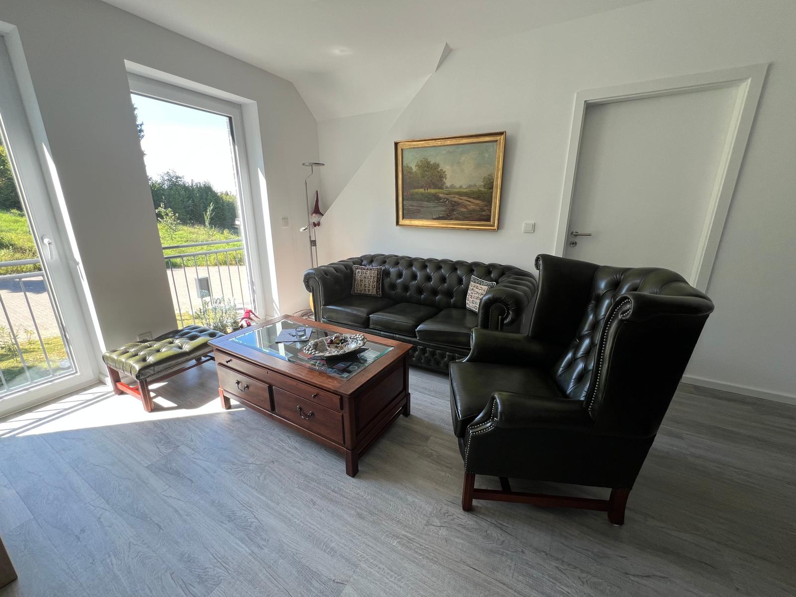 Wohnzimmer mit Ledersofa, Sessel und Glas-Tisch. Balkon mit Blick auf Grünfläche.