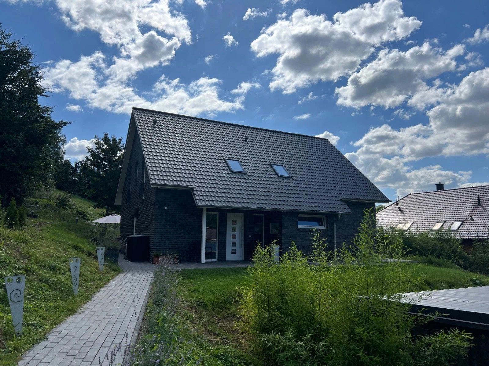 Ein modernes Haus mit dunklem Dach und weißen Fensterrahmen. Ein gepflasterter Weg führt zum Eingang.