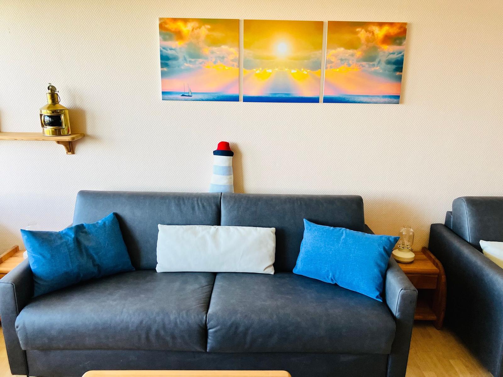 Ein grauer Sofa mit blauen und weißen Kissen. Wandbild zeigt Sonnenuntergang mit Segelboot.