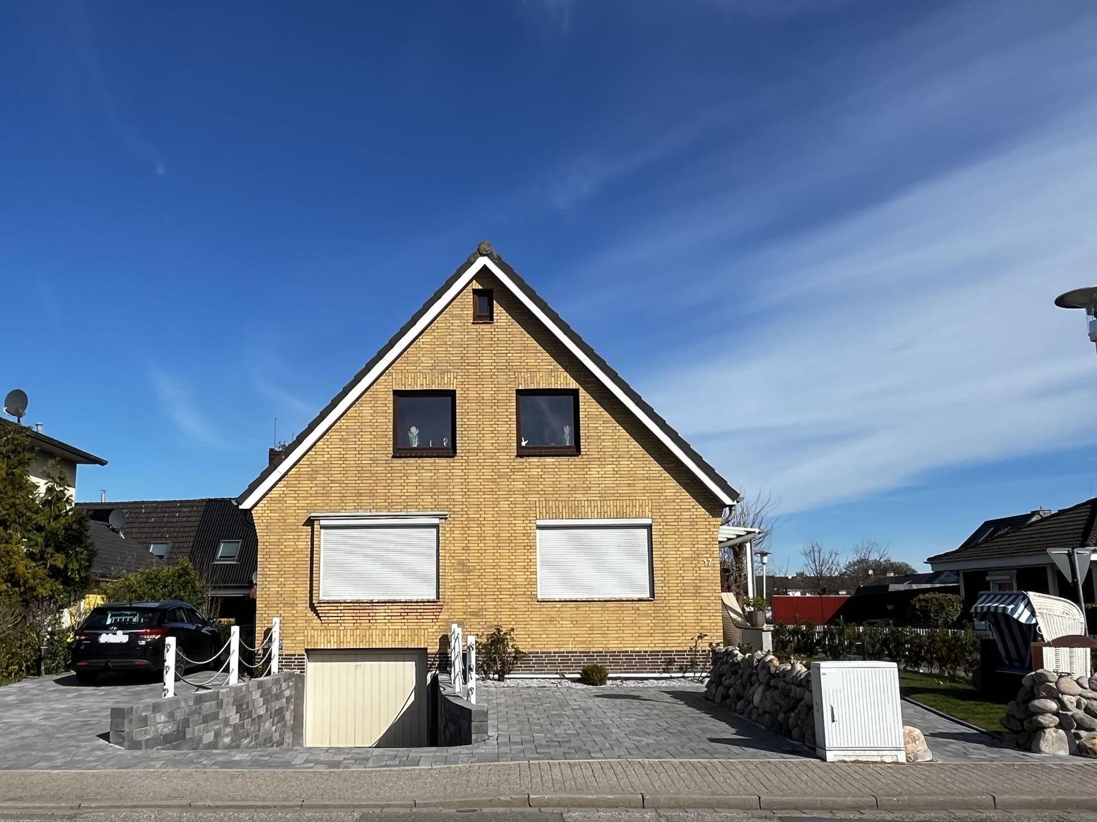 Ein gelbes Haus mit Dachgauben und Rollläden. Vorhof mit Steinmauer und Parkplatz.