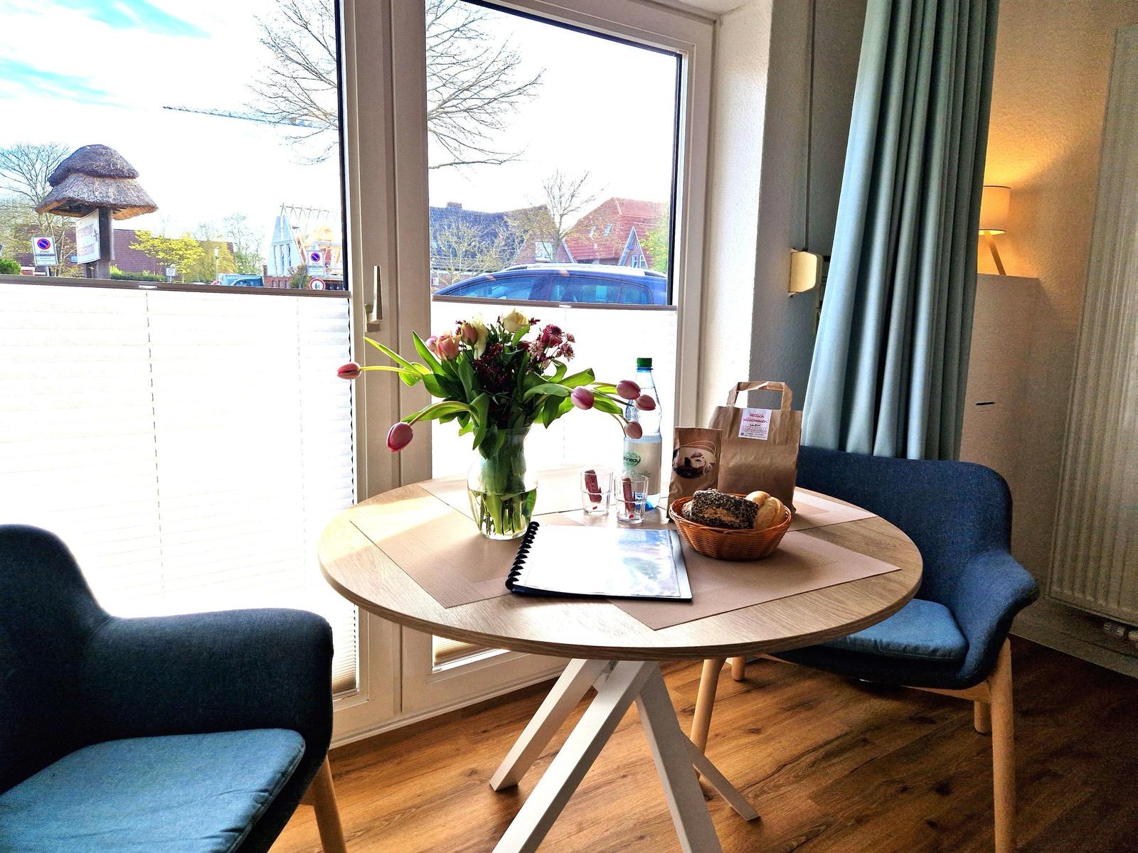 Rundtisch mit Blumen, Brot und Getränk vor großem Fenster.