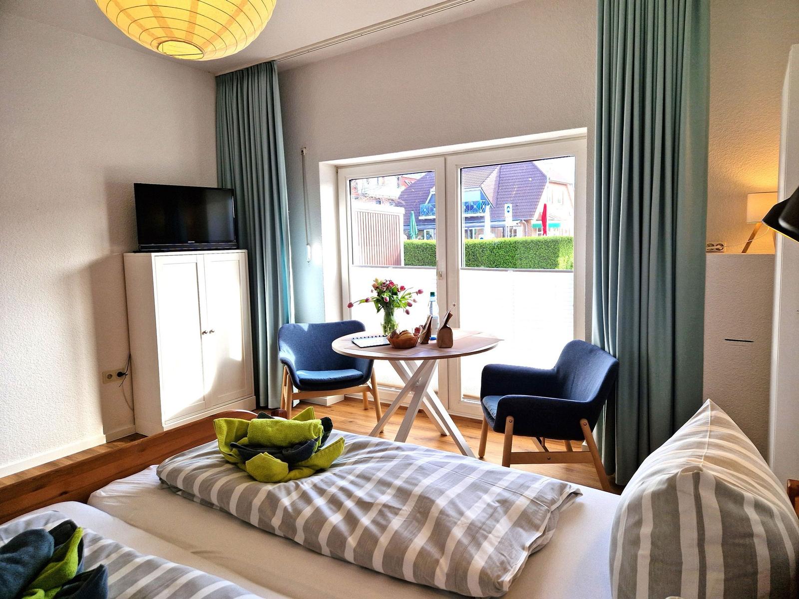 Ein Schlafzimmer mit Bett, TV, Tisch und Stühlen. Durchgang zum Balkon mit Blick auf die Straße.