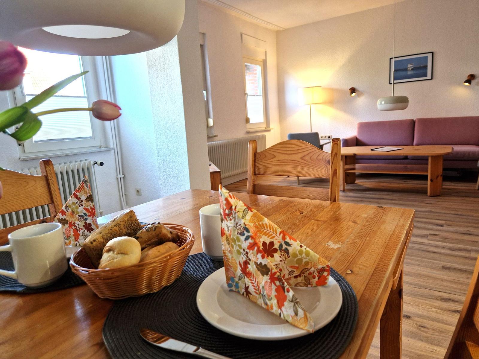 Dining room with wooden table, chairs, and basket of bread. Living room with sofa in background.