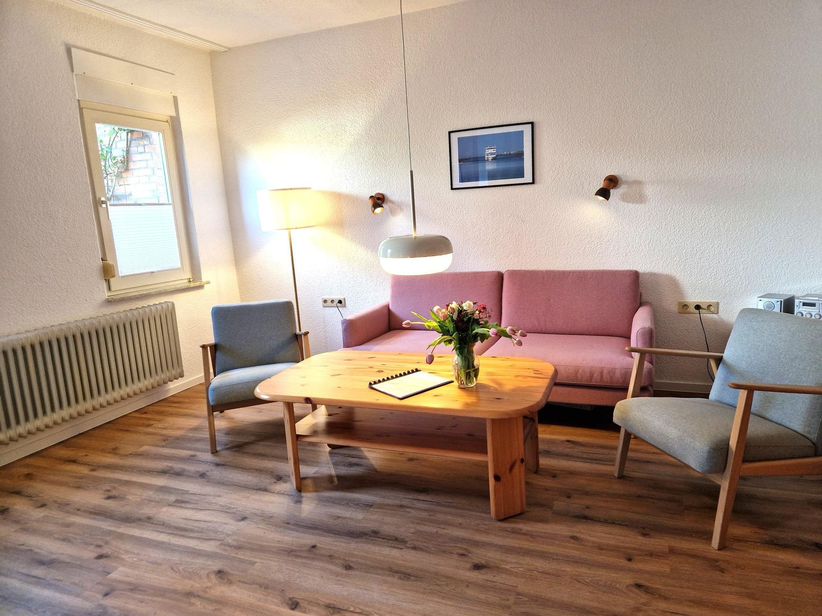 Living room with pink sofa, two chairs, and wooden table. Window, radiator, and wall lamps visible.