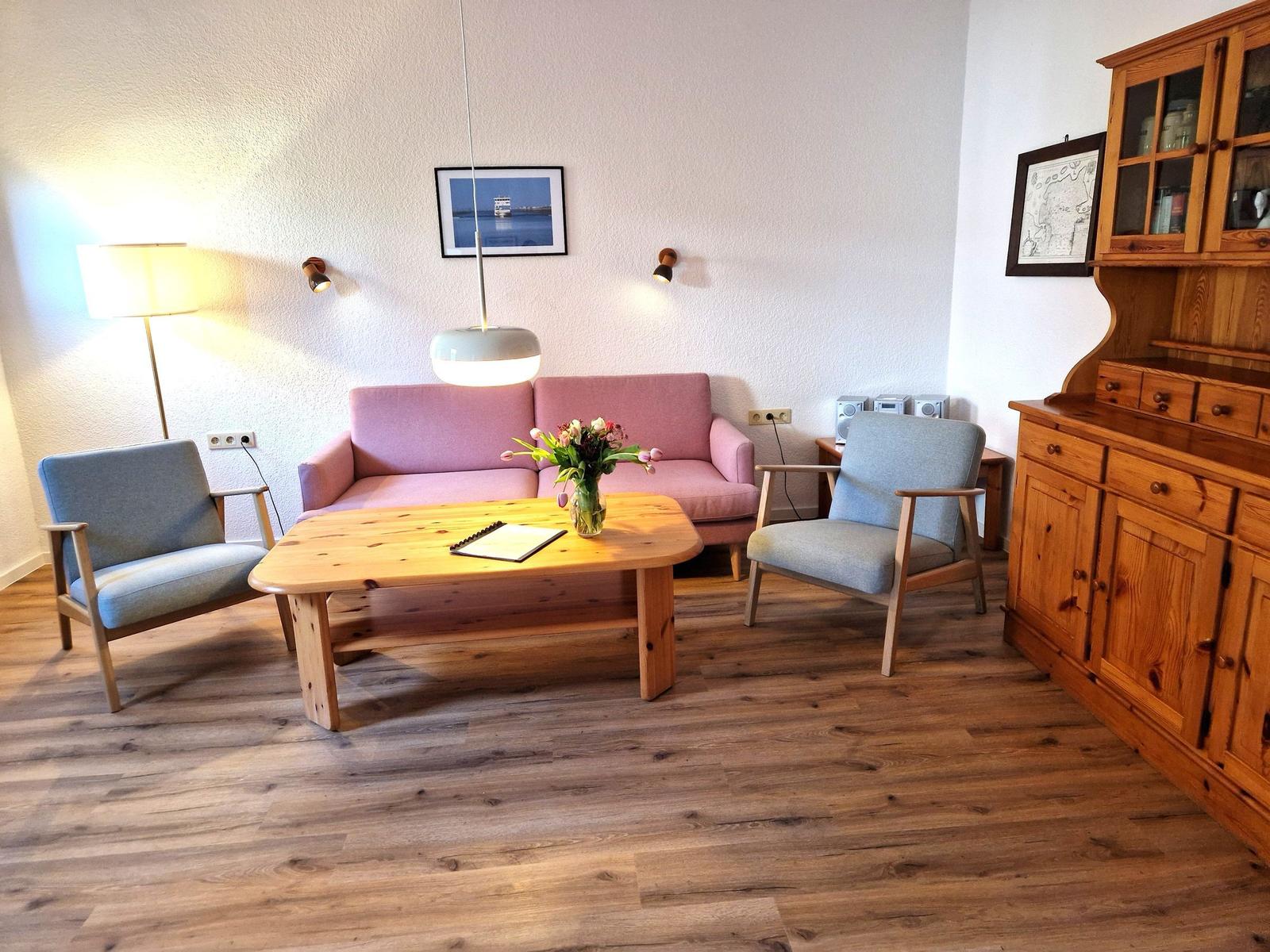 Living room with pink sofa, two blue chairs, and wooden table. Wooden cabinet and wall pictures are visible.