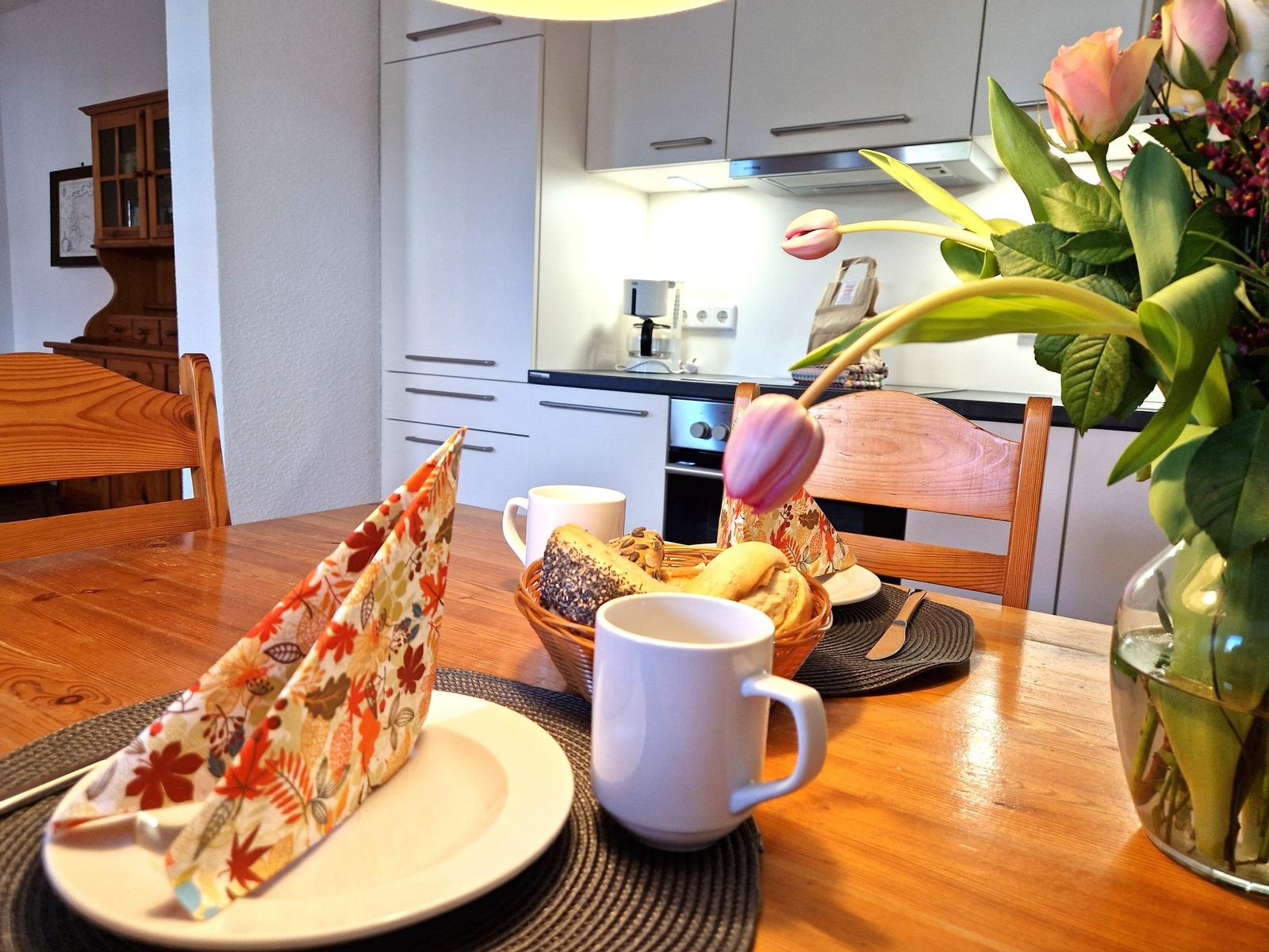 Wooden table with mugs, bread, and flowers in dining area.