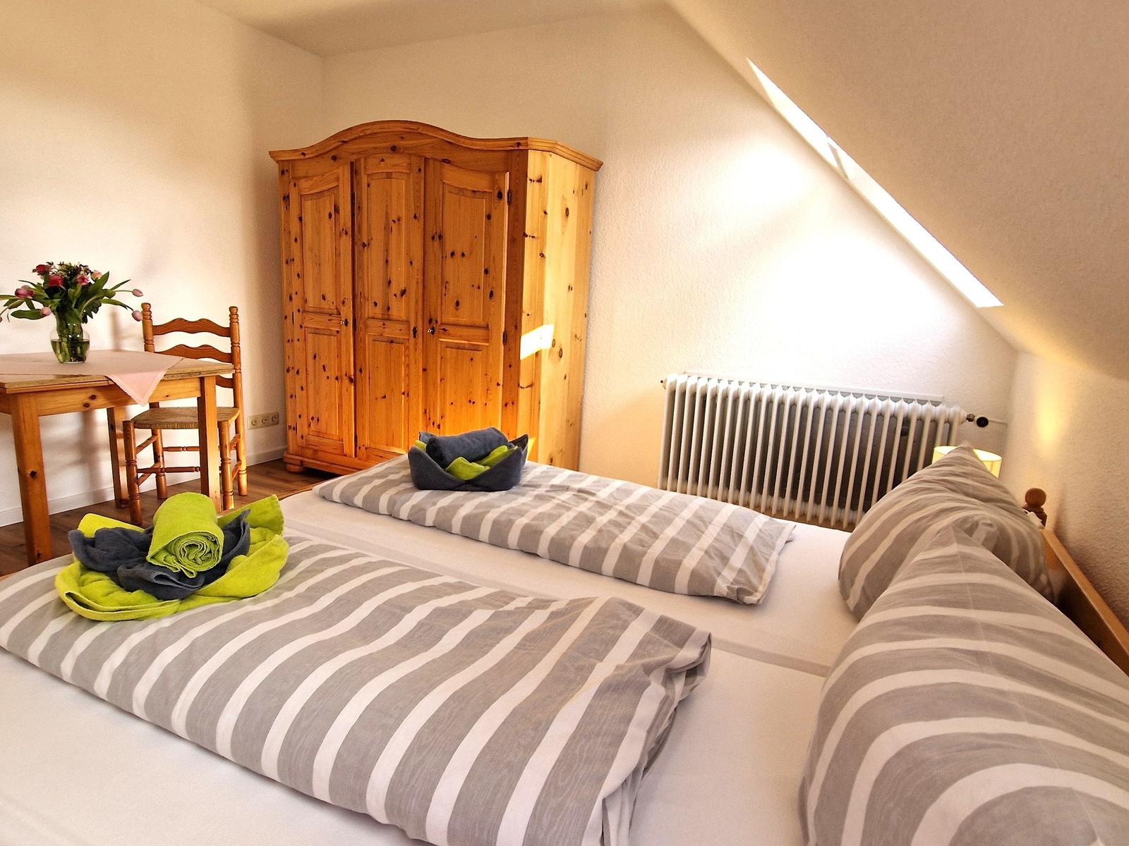 Zimmer mit Doppelbett, Holzschrank, Tisch und Stuhl sowie Heizung unter Dachschräge.