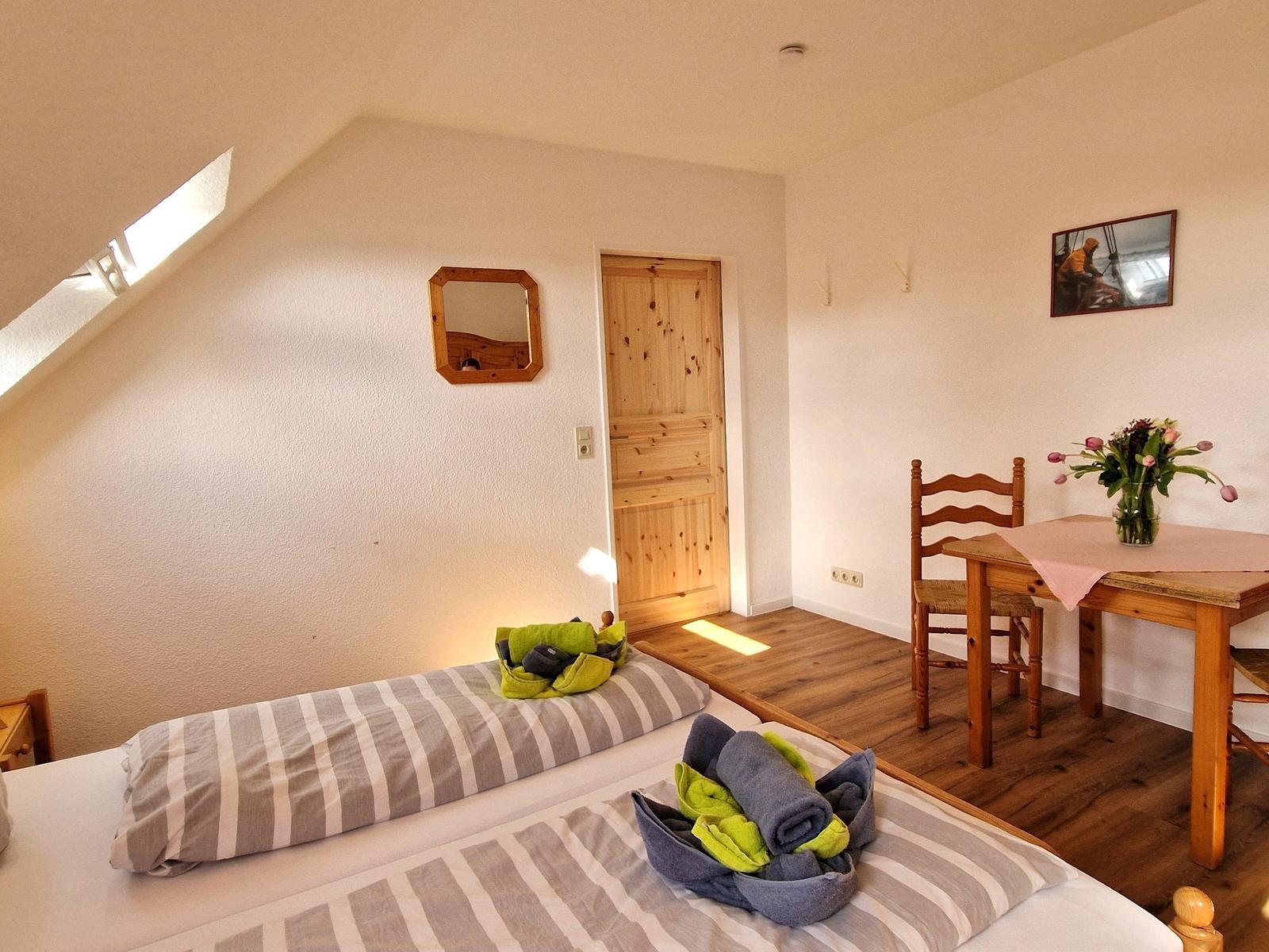 Zimmer mit Dachschrägen, Bett, Tisch und Stuhl. Holztür und Spiegel an der Wand.