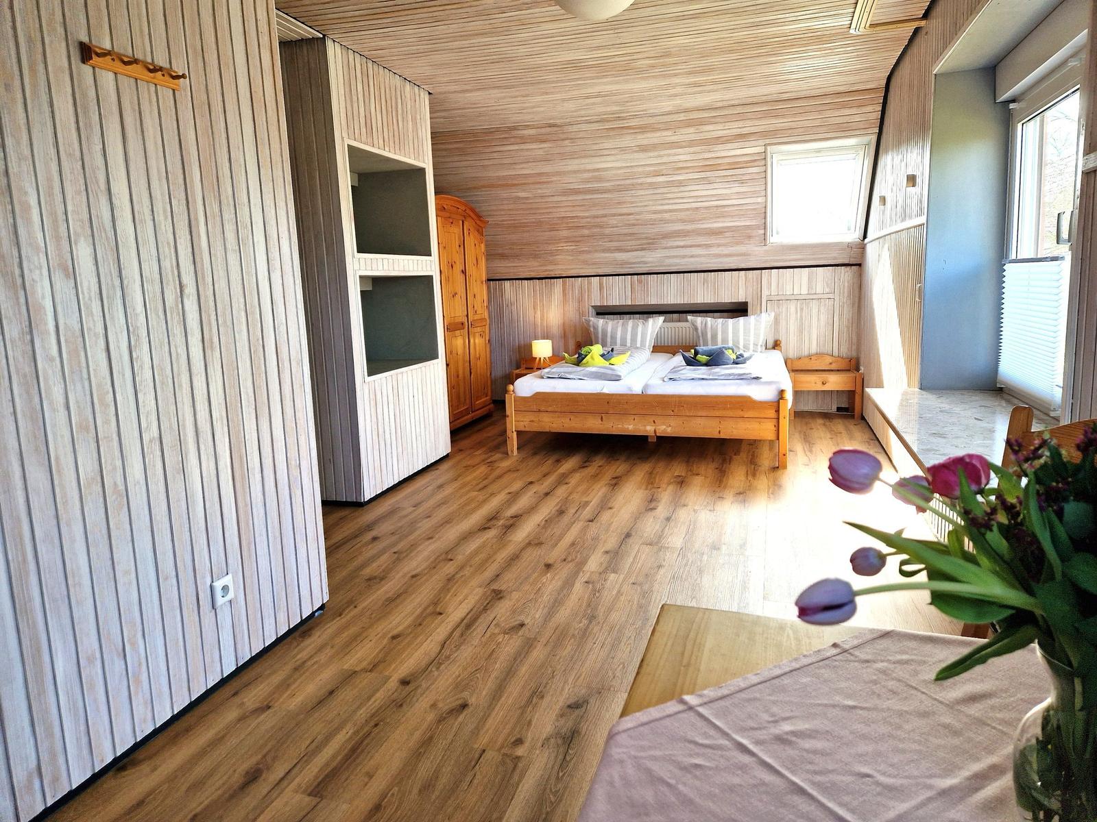 Schlafzimmer mit Holzboden, Bett und Fenster. Vase mit Tulpen im Vordergrund.